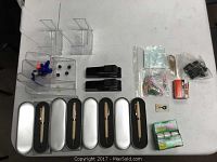 Photo showing five wood pens in individual metal gift boxes, two black classic staplers, clear acrylic display organizers containing paper clips, colored pins, push pins, rubber bands and other small office accessories, as well as packs of tape and stapler pins.