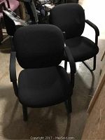 Two black office chairs with cushioned seats and backs, black armrests and metal legs shown in an indoor setting.