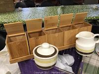 Four wooden tea bag holders with lids arranged on a matching wooden tray, accompanied by a ceramic milk pitcher and sugar bowl with lid, featuring brown and yellow stripes.