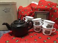 Brown ceramic teapot with lid, six white floral coffee cups, red cloth background, large cream crock partially visible