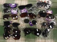 Overview photo showing 16 pairs of sunglasses arranged on a table, some with tags and in plastic packaging.