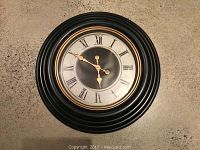 Front view of large round wall clock with black concentric ridged frame and Roman numeral dial.