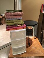 Metal bistro table with round top partially visible behind stack of towels and plastic drawer unit placed on a wooden table.