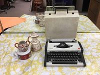 Photo showing Olympia manual typewriter with black keys alongside hand painted ceramic items on a floral tablecloth.