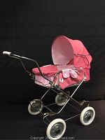 Full side view of vintage pink fabric doll pram with chrome frame and white wheels, black backdrop