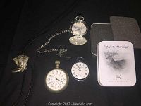 Full view of lot items including three pocket watches with one open showing interior cover, chain, exclusive edition tin box, and cowboy boot neck tie.