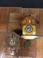 Overview of both cuckoo clocks side by side on wood surface showing size and design differences