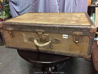 Front view of rectangular leather suitcase with rope handle and metal latches, showing A.T.C. stamped on front and aged wear.
