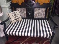 Front view of antique black wooden porch bench with black and white striped seat upholstery and decorative cushion on the left side.