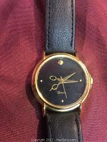Front view of quartz wristwatch with scissors design on black dial and gold-tone case with black leather band.