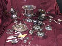 Overview of silver plated tea and coffee pots, champagne bucket, cream and sugar set, candlesticks, serving tray, and assorted flatware on a burgundy fabric backdrop.
