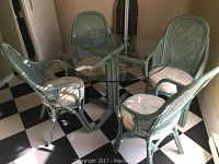 Full set photo showing sea foam colored glass top table with rattan pedestal base and four matching rattan cane back chairs with loose upholstered seats on a black and white checkered floor.