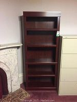 Front view of the dark wood finish bookcase showing five shelves and decorative grooved top panel next to a fireplace and filing cabinet.