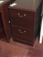 Front view of filing cabinet showing two drawers and brass handles, wood finish, placed on carpet.