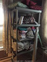 Full view of grey plastic shelving unit filled with various linens including folded towels, blankets, fabrics, and a comforter on top shelf