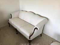 Front and side angle showing full length of cream-colored settee with wooden armrest accents and three seat cushions on carpeted floor against plain wall.