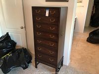 Tall wooden chest with six drawers, brass handles, positioned between door and wall with black bags nearby.