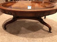 Front angle view of oval coffee table showing leather top, decorative gold edge, drawer front and pedestal base