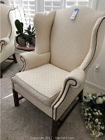 Front-right angle view of one cream-colored upholstered wingback chair showing patterned fabric, nailhead trim, removable seat cushion, wooden legs.