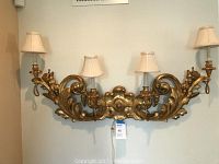 Full frontal image of the antique four lamp wall light fixture mounted on a wall with four beige fabric lampshades and gold leaf ornate carved wood frame.