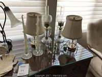 Photo of two small lamps with shades, two crystal hurricane lamps with glass and metal parts with one hanging crystal detached, clear glass candleholders with white candles, and two small vintage perfume bottles on a wooden desk.