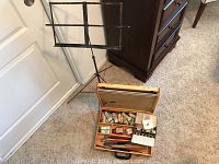 Photo showing a vintage metal music stand and a wooden art box on a carpet with the box open revealing painting supplies.