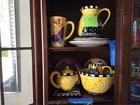 Photo showing two mugs, a pitcher, two plates stacked, all with colorful Laurel Burch style cat designs