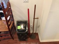 Photo of antique black metal coal scuttle and wooden washing agitator side by side with a red ruler for scale, next to wooden chairs and wall outlet.