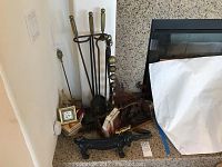 Full view of the fireplace area showing the iron dog boot scraper in the foreground, fireplace tools with brass handles in the stand, brass bellows, and miscellaneous small items grouped together.