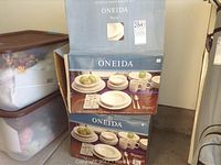 Photo showing two boxed sets of Oneida Regency 32-piece dinnerware stacked vertically with other storage boxes nearby.