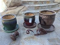 Front view of three glazed clay planters on trays along with metal bird sculptures, metal snail, and curled metal scrolls placed on stone steps.