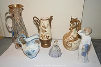 Full view of six items including clear glass pitcher, two cream porcelain pitchers, blue and white vase, female figurine in lavender dress, and Lladro figurine