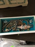 Top drawer of the jewelry box with various costume jewelry and pins including a beaded necklace, porcelain doll pendant, and earrings.