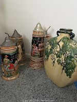 Photo of two German beer steins with lidded pewter tops and colorful beer hall themed designs, along with a beige and green pottery vase with grapevine detailing.