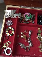 Open rectangular wooden jewelry box with compartments and red velvet lining showing assorted pins, brooches, rings, bracelets including horse-drawn wagon pin and colorful spiral brooches.