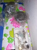 Sterling silver lidded glass jar with floral embossed lid, and small baby spoons in plastic wrapping on a colorful floral background.