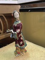 Asian porcelain figurine of a woman in traditional dress, detailed with floral designs, beside wood furniture and kitchen appliance.