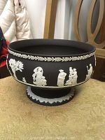 Front view of black Wedgewood pedestal bowl with white classical relief figures and foliage around the exterior.