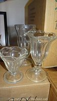 Three different sizes of clear fluted soda fountain glasses shown on stacked cardboard boxes, indicating sets of 12 per box.