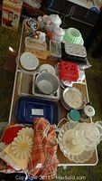 Overview of various vintage kitchen bakeware and linens laid out on tables, includes Pyrex and ceramic bakeware, spice tins, several plastic containers, and linens.