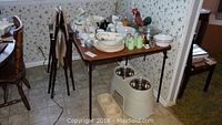 Wide view of card table holding various vintage kitchenware and china, with folding chairs stored next to it.