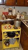 Photo showing the yellow 1950s metal kitchen cart holding cast iron skillets, a metal whistling tea kettle, metal steamer pot, and various kitchen items including decorative mugs and ceramic pieces.