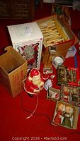 Photo showing lighted Santa figure with original cardboard box and assorted Christmas decorations arranged on red carpeted floor.