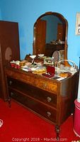 Vintage wooden dresser with three drawers, attached arched mirror, some items displayed on top, wood grain and drawer details visible, situated against blue wall on red carpet.