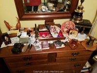 Vintage masculine items spread on wooden dresser including binoculars, cowboy clocks, metal box, lamp, figurines and various accessories.