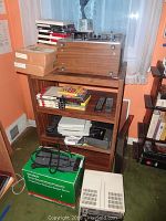 Full view of wooden shelving unit holding Aka I reel to reel player atop, boxes and reel to reel tapes on shelves, Hanimex SR8600 projector box and other electronics on floor in front