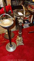 Two floor standing ashtrays placed next to each other; one ornate metal with pedestal base and one black with silver base and handle.