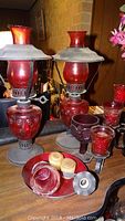 Two matching red glass oil lamps with metal tops and bases, additional red glassware including Avon cranberry glass, a candle holder, and a dish with lids.
