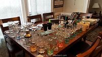 Wide view of dining table with various glassware items, cocktail shaker, kitchenware including knives and pots in the background.