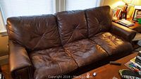 Front view of a brown leather three-seat couch showing wear and creasing on cushions and armrests, with metal upholstery tacks visible along the edges.